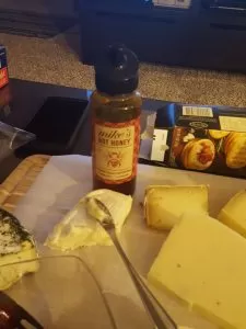 A close-up shot of a rustic cheeseboard featuring a wedge of blue cheese, slices of cured ham, and a small pot of hot honey, showcasing a gourmet spicy and sweet pepper combination.